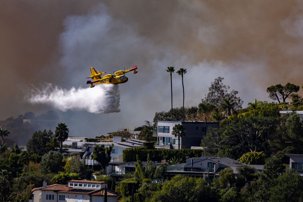 Yellow and red firefighting aircraft drops water over a hillside neighborhood as thick wildfire smoke fills the sky behind homes and palm trees.