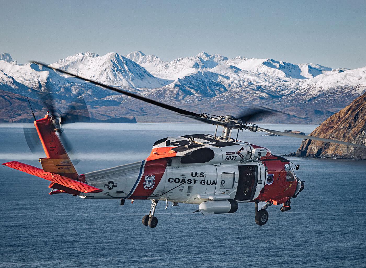 U.S. Coast Guard rescue helicopter flying over Alaska coastline with a snowy mountain range in the background