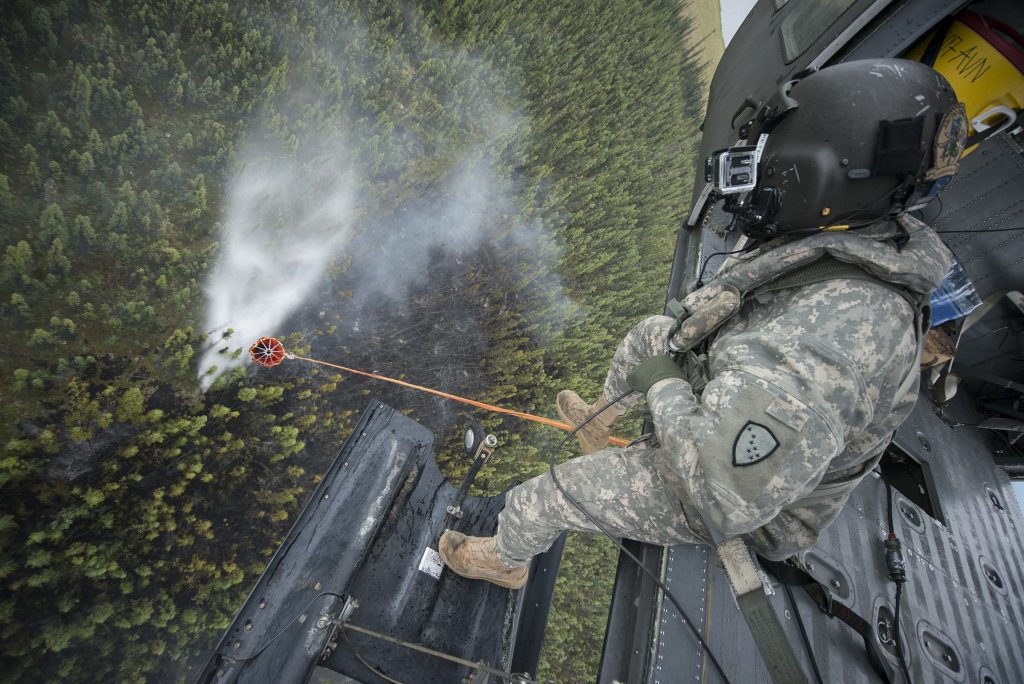 An Alaska Army National Guard crew member leans from a helicopter as a suspended bucket releases water over burned forest.