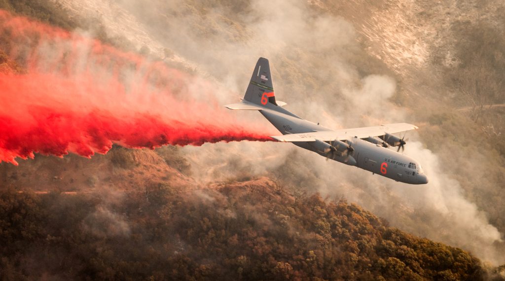 A C-130J drops a long red line of fire retardant across a smoky, rugged hillside.