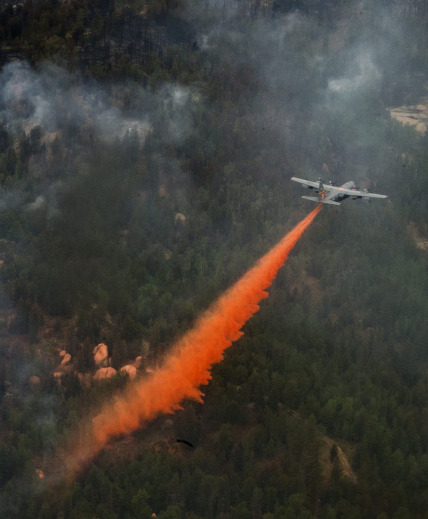 A C-130 aircraft drops a long orange line of fire retardant over smoky, forested terrain.