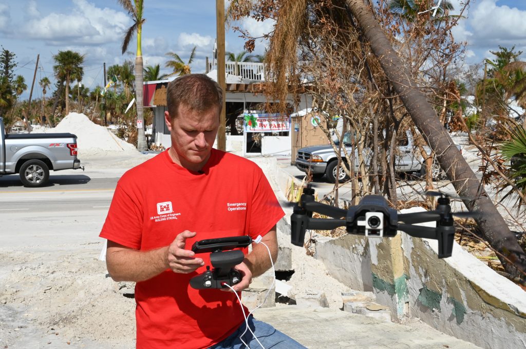A man in a red shirt holds a drone controller as a small drone hovers beside him on a storm-damaged street.