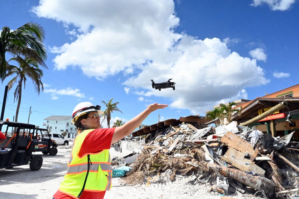 A drone pilot in a hard hat and safety vest launches a drone above hurricane debris on a Florida beach.