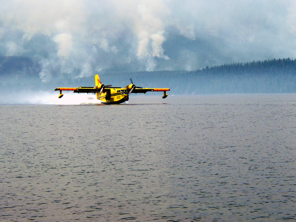 A yellow Canadair CL-215 skims the surface of a lake with smoke rising from forested hills behind it.
