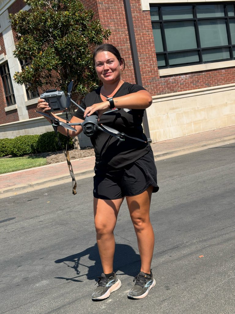 Courtney, a drone pilot, smiles on a brick-lined Waco street holding her drone and controller.
